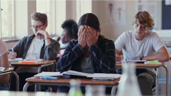 Shows a classroom, with students taking a test and one looking stressed, with her head in her hands