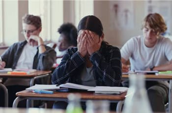 Shows a classroom, with students taking a test and one looking stressed, with her head in her hands